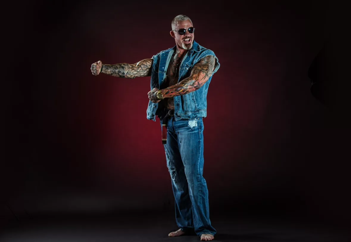 Rick Koenig, dramatic studio portrait in denim vest and sunglasses on a dark maroon-lit backdrop